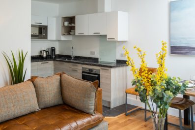 open living space with modern kitchen and brown leather sofa with tweed style cushions and vase of yellow flowers