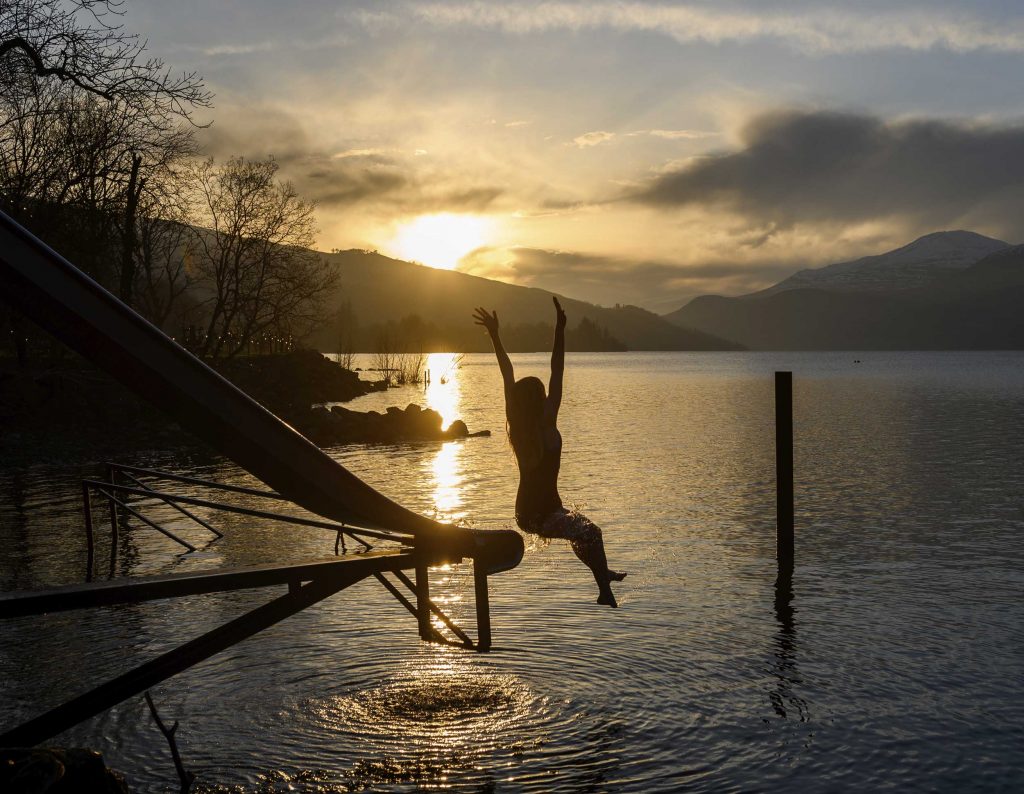 Silhouette of young woman jumping off a slide into the Loch with a golden sunset and mountain in background